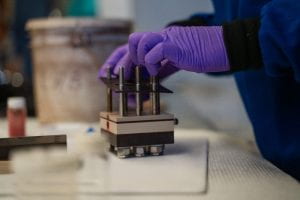 a researcher assembles a cell used to produce iron metal from iron oxide. the cell is a stack of plastic sheets and gaskets held together by large bolts.